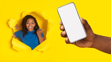 Smartphone Boş Ekranı 'nı gösteren mutlu siyah kadın, Büyük Mobil Uygulamayı Öneriyor ya da Stüdyodaki Yırtık Sarı Kağıt Deliğinde Poz Vermeyi Öneriyor. Telefonun için harika bir uygulama. Panorama