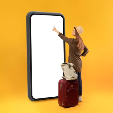 Seyahat uygulaması. Bayan Gezgin, Stüdyodaki sarı arka planda sırt çantası ve bavul pozu ile boş ekrana dokunarak büyük bir telefon kullanıyor. Tatil ve Turizm Konsepti. Kare Vuruş