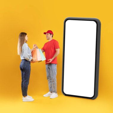 Mobil Alışveriş. Teslimatçı, sarı arka planda boş ekranı olan büyük cep telefonlu bir kadına alışveriş torbası veriyor. E-Ticaret. Ticaret ve Teslimat Başvurusu. Kare