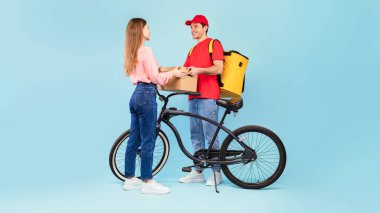 Teslimat servisi. Bisikletli ve sırt çantalı kadınlara paket kutusu teslim eden erkek kurye stüdyoda mavi arka planda duruyor. Tam Uzunluk Çekimi, Panorama