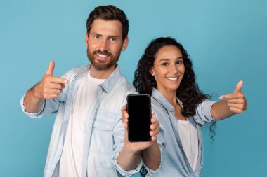 Gülümseyen Avrupalı genç adam ve Arap kadın parmaklarını ekranı boş, mavi arka planda, stüdyoda izole edilmiş, kapalı akıllı telefondan gösteriyor. Yeni uygulama, aygıt, kazanç, harika teklif, reklam ve satış sunumu