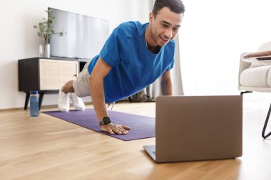 Spor kıyafetli genç bir orta doğulu şınav çekiyor, bilgisayarında ev bilgisayarında, oturma odasında iç dizüstü bilgisayar var. Sportif adam günlük ev eğitiminden zevk alıyor.