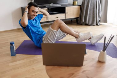 Spor giyimli aktif genç bir orta doğulu adam evde egzersiz yapıyor, karın kasları üzerinde çalışıyor, dizüstü bilgisayara bakıyor ve gülümsüyor, online fitness videosundan keyif alıyor, yer kopyalıyor.
