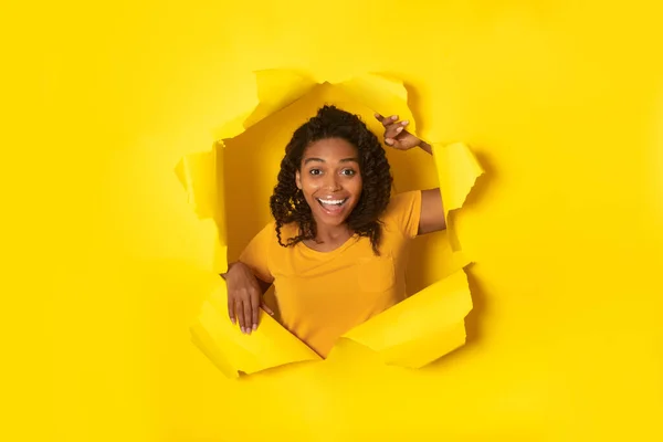 Joyful Black Lady Posing Tearing Paper And Looking Through Hole In Yellow Studio Background, Advertising Wow Offer Smiling To Camera. Commercial Advertisement Banner