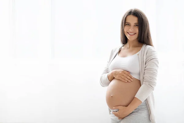 年轻的孕妇穿着舒适的家居服 站在白色的背景上笑着 抱着她的大肚子 等着孩子 带着复制空间的全景 母性概念 — 图库照片