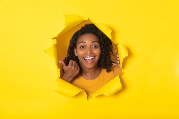 Excited Black Female Posing Looking Through Hole In Torn Paper Pointing Thumbs At Yellow Background. Studio Shot Of Lady Advertising Great Offer Smiling To Camera. Advertisement Banner Concept