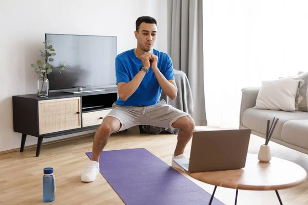 穿着运动服的年轻的阿拉伯运动员蹲在客厅的内部 在笔记本电脑上观看在线健身课 Covid 19期间 健身博客 器械和家庭锻炼 — 图库照片