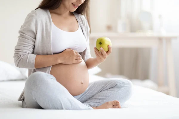ホームウェアの認識できない長い髪の若い妊婦の自宅でベッドの上に座って 大きなおなかに触れ コピースペースと新鮮な緑のリンゴ パノラマを保持する 妊娠中の健康的な食事 — ストック写真