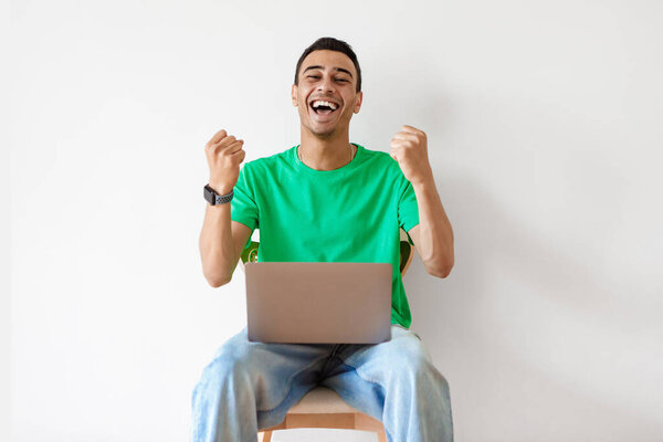 Online win concept. Emotional arab guy celebrating success with laptop, sitting on chair at home, man raising hands and exclaiming with excitement, copy space