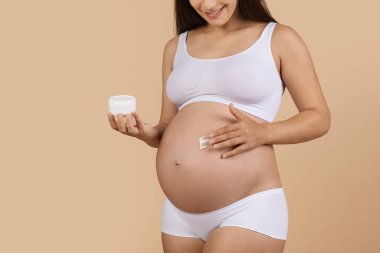 Beyaz iç çamaşırlı, tanınmayan hamile bir kadın, bej renkli stüdyo arkaplanına poz verirken vücuduna losyon sürüyor. Beklenmedik kadın gerilme izlerine karşı tedavi görüyor.