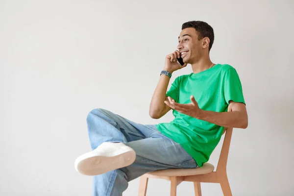 Excited arab guy speaking on cellphone, sitting on chair over white wall, having phone conversation, copy space. Middle eastern man communicating online on smartphone