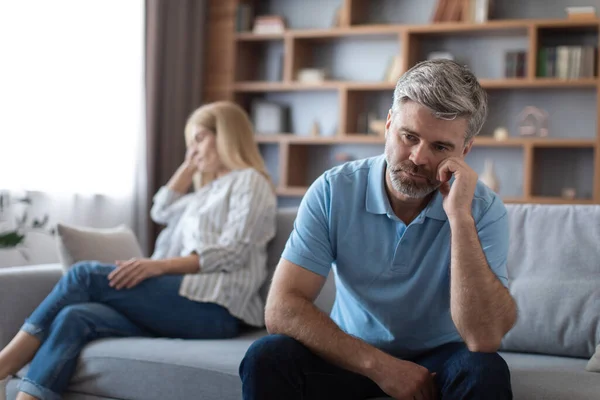 Boring sad mature caucasian male ignoring lady after quarrel and thinking about breaking up on sofa in living room interior. Divorce, relationship problems and depression at home during covid-19