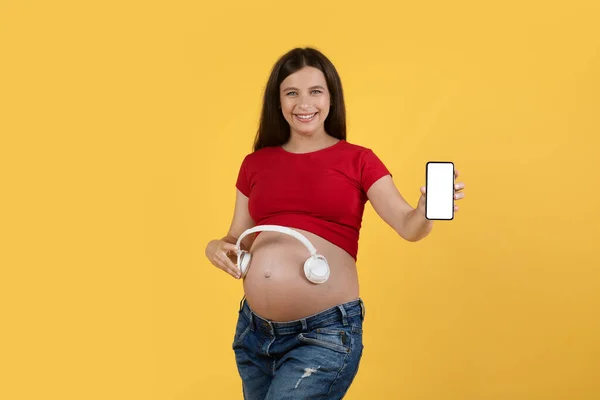 妊娠中の幸せな女性は腹の近くにワイヤレスヘッドフォンを保持し カメラでブランクスマートフォンを実証 子宮の中で赤ちゃんのための音楽をお勧めします陽気な若い期待の女性 モックアップ — ストック写真