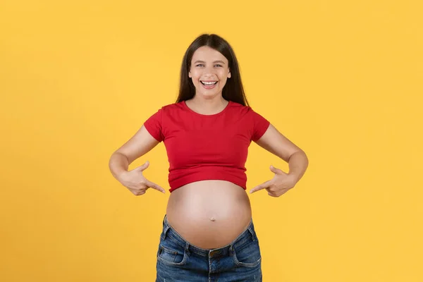 2本の指で彼女の大きな妊娠腹を指している陽気な若い女性 おなかを示す積極的な期待の女性 健康的な妊娠を楽しんで 今後の母親 黄色の背景 コピースペース — ストック写真