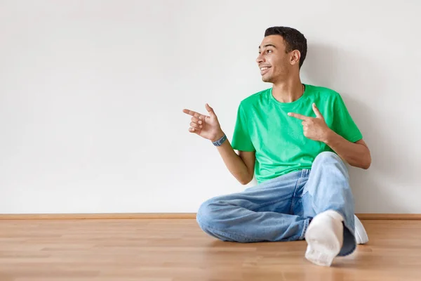 酷广告 年轻的阿拉伯男子坐在地板上 把空余的空间指向白墙 积极的家伙展示广告或弹药的位置 — 图库照片