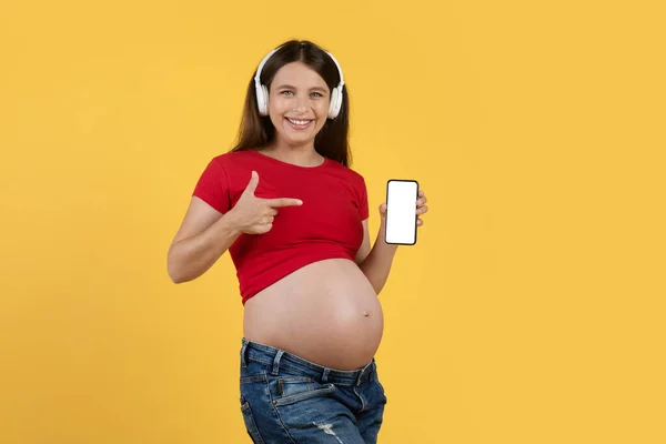 手に空白のスマートフォンを指している無線電話で幸せな妊娠中の女性 笑顔期待の女性は 新しい音楽アプリをお勧めします 黄色のスタジオの背景に孤立立ちます モックアップ — ストック写真