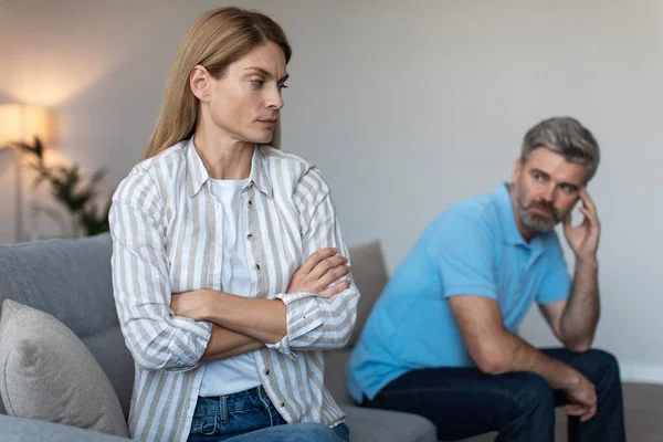 Offended mature caucasian husband ignoring wife after quarrel and thinking about divorce on sofa in living room interior. Break up, relationship problems, scandal at home during covid-19 outbreak