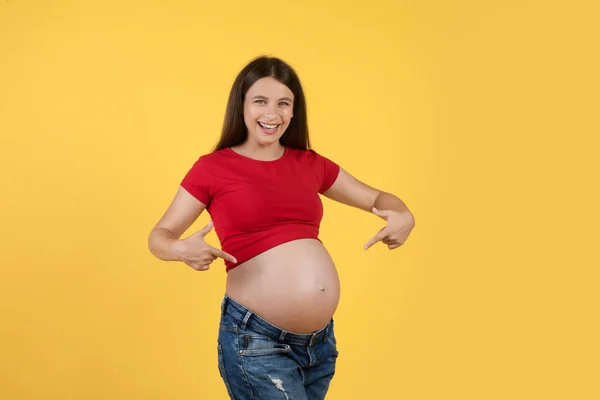 明るい妊娠中の女性は黄色の背景に隔離されている間 彼女の腹で指を指し カメラで笑顔 彼女の大きなおもちゃを示す幸せな若い期待の女性 妊娠時間を楽しんで — ストック写真