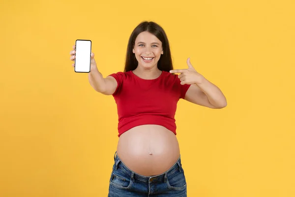 新しいアプリだ 朗らかな妊娠中の女性が空白のスマートフォンを表示し それを指して 幸せな笑顔期待の女性妊娠追跡のためのモバイルアプリケーションをお勧めします 黄色の背景 モックアップ — ストック写真