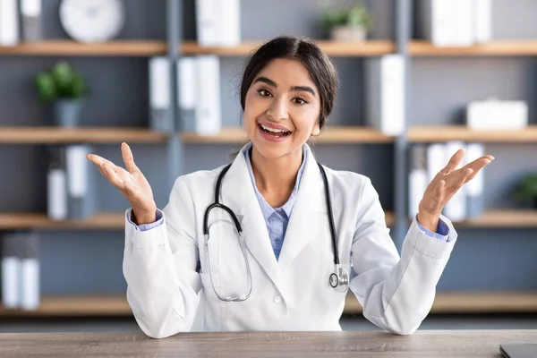 Smiling millennial attractive indian woman doctor in white coat gesturing and talking at workplace in clinic office interior. Medical consultation remotely, new normal, social distancing due covid-19