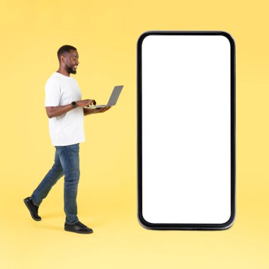 Siyah erkek, sarı arka planda boş ekranlı, büyük telefonun yanında dizüstü bilgisayar kullanıyor. Serbest Çalışan Adamın Stüdyo Çekimi Bilgisayar ve Mobil Uygulama Reklamları 'nda çalışıyor. Kare, Mockup