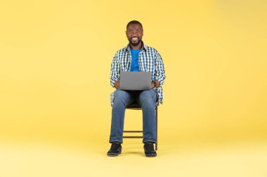 Neşeli Siyah Adam internette dizüstü bilgisayar kullanıyor ve internet reklamını yapıyor sarı stüdyo arka planında oturuyor, kameraya gülümsüyor. Bilişim kariyeri konsepti. Tam Uzunluk Çekimi