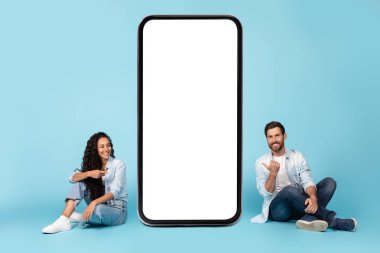 Neşeli bin yıllık beyaz erkek ve orta doğulu bayan yerde oturur, boş ekranlı akıllı telefonu işaret eder, mavi arka planda ve stüdyoda izole olur. İş ve blog için yeni uygulama, reklam ve teklif