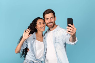 Sakallı neşeli beyaz bir adam Arap bir bayana sarılıyor, el sallıyor, akıllı telefondan video görüşmesi yapıyor, mavi arka planda, stüdyoda izole edilmiş. Uzaktan görüşme, harika reklam ve teklif, yeni normal, covid-19