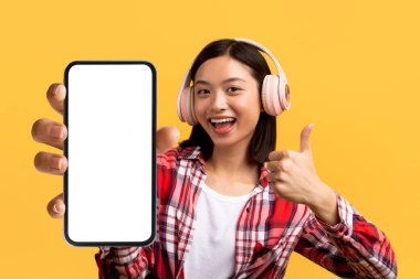 Güzel müzik uygulaması. Heyecanlı Asyalı kadın boş cep telefonu ekranı gösteriyor ve sarı stüdyo arka planında kulaklık takıyor. Başvuru ya da web sitesi reklamı için model