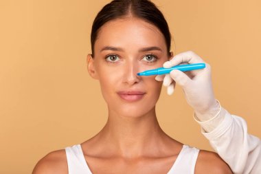 Rhinoplasty concept. Doctor making marks on patients face, young caucasian lady on consultation at surgeon, standing on beige background. Facial plastic surgery concept clipart