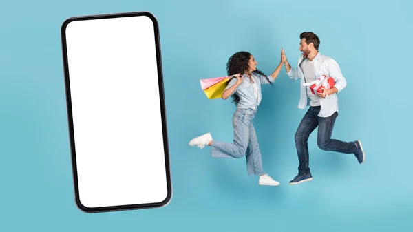 Smiling excited millennial caucasian guy and arab female with gift and shopping bags jump in air, give high five near huge smartphone with blank screen, on blue background. Fun, success, win and sale