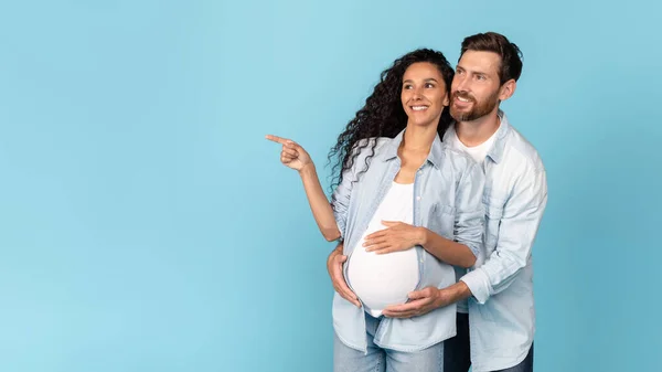 Cheerful millennial european male with beard hug pregnant arab lady with belly, lady point finger at empty space isolated on blue background. Great ad and offer, couple enjoy love, expecting baby