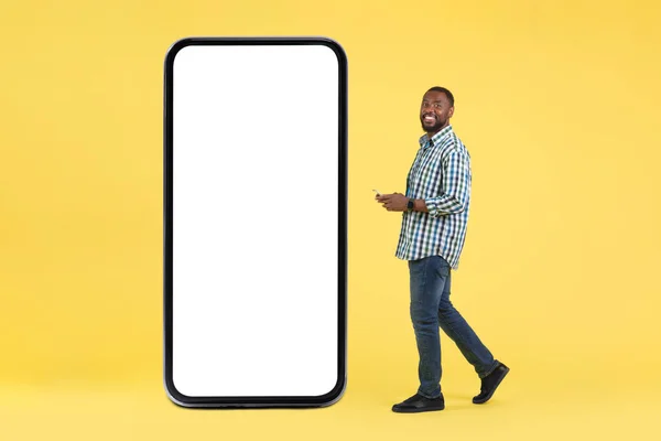非裔美国男性使用智能手机发短信靠近大手机与空屏幕站在黄色工作室背景 移动应用程序概念 — 图库照片