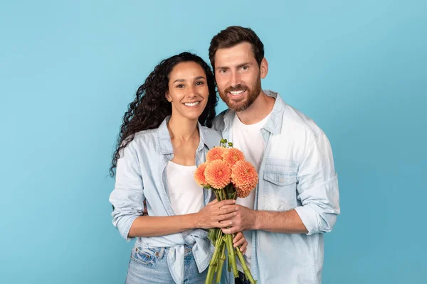 令人欣喜的是 千禧年的阿拉伯女子和欧洲男子留着胡子 看着相机 手持鲜花 与蓝色背景隔离 工作室 广告和提议 生日庆祝 爱情和浪漫 — 图库照片