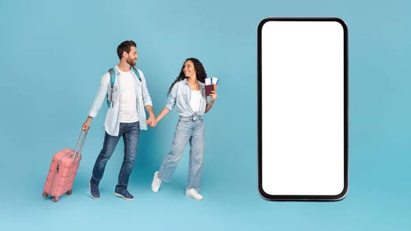 Smiling young caucasian male and arabic female with suitcase and air tickets hold hands, go to smartphone with blank screen, isolated on blue background. Ad and offer, fun, vacation and travel app