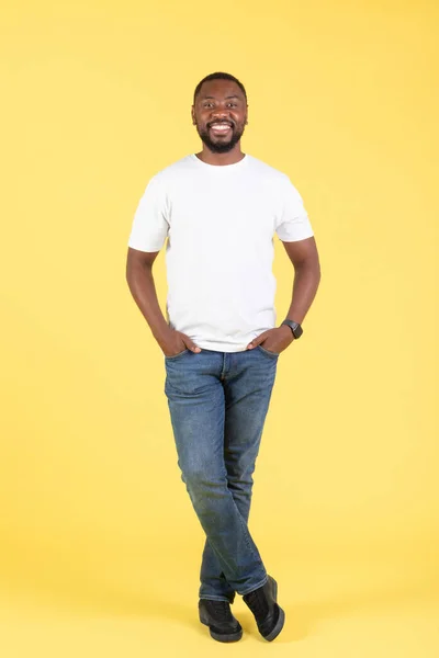 Handsome Black Male Smiling Looking At Camera Posing Standing Over Yellow Studio Background, Wearing T-Shirt And Jeans. Great Offer Concept. Vertical, Full Length Shot