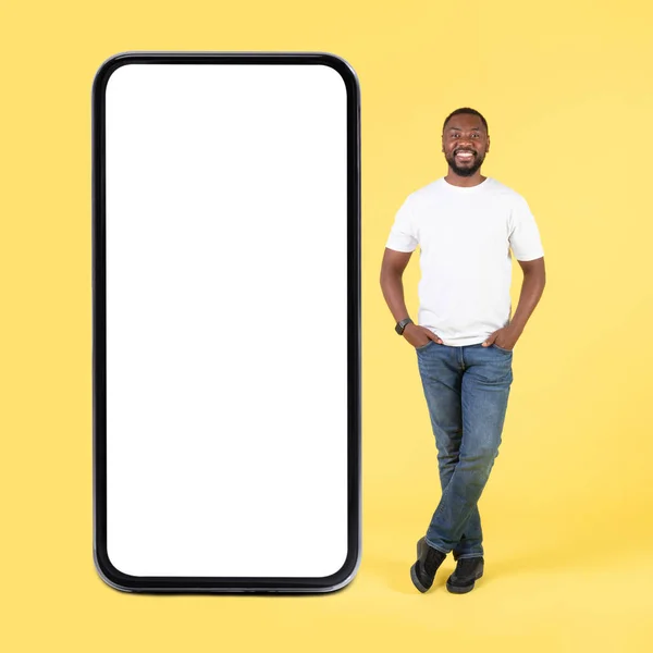 快乐的非洲裔美国人 站在大智能手机旁边 手牵手插在口袋里 在黄色背景下发布新的移动应用程序 Studio Shot Mockup — 图库照片