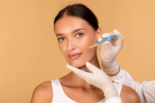 Lip augmentation concept. Doctor hands in gloves with syringe near female face, happy woman receiving beauty injection, standing over beige studio background
