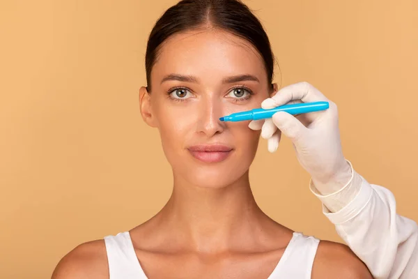Rhinoplasty concept. Doctor making marks on patients face, young caucasian lady on consultation at surgeon, standing on beige background. Facial plastic surgery concept