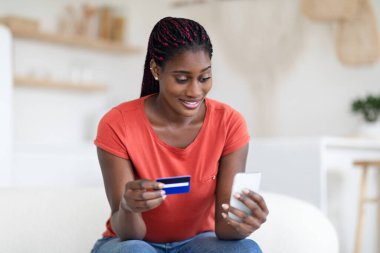 Zeki telefon ve kredi kartıyla online alışveriş yapan gülümseyen Afrikalı Amerikalı kadın, oturma odasında dinlenirken internetten alışveriş yapıyor.