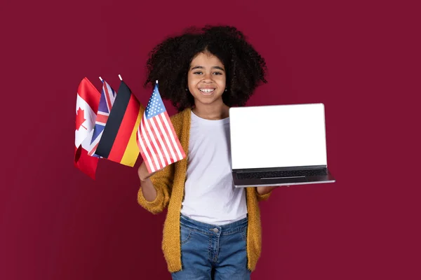 快乐漂亮的黑人小女孩 留着浓密的头发 手里拿着笔记本电脑 上面有白色的空白屏幕 挂着来自不同国家的国旗 有勃艮第的工作室背景 还有模型 外语在线学校 — 图库照片