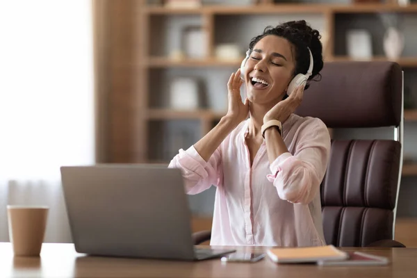 Stress relief at workplace concept. Happy young businesswoman listening to music and singing, lady sitting at workdesk in front of computer, using wireless headset, copy space