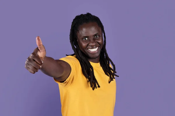 黄色のTシャツを着た前向きなハンサムなアフリカ系アメリカ人の長髪の若い男が親指を見せ 紫色のスタジオの背景にカメラで笑みを浮かべて 何かを推薦し スペースをコピーします ジェスチャーコンセプト — ストック写真