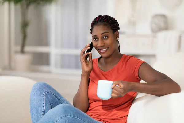 Domestic Leisure. Happy Young Black Woman Talking On Phone And Drinking Coffee While Relaxing On Couch At Home, Smiling African American Female Resting On Comfortable Sofa In Living Room