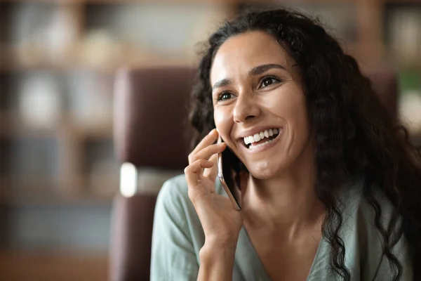Closeup of happy pretty young woman in casual outfit sitting at table at home office, having conversation on phone and smiling, talking with business partner, looking at copy space
