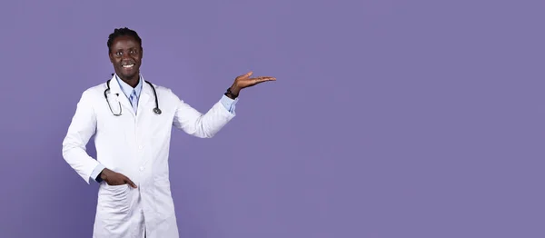 広告のためのコピースペースを指して 紫色のスタジオの背景 パノラマの上に笑みを浮かべて聴診器を持つ作業服の医者で陽気なハンサムな長髪の若い黒人男性 医療の概念 — ストック写真