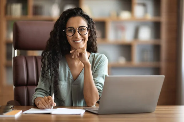 Positive brunette millennial woman in eyeglasses using laptop at office, lady sitting at workdesk in front of computer, smiling at camera, copy space. Career, business opportunities for women