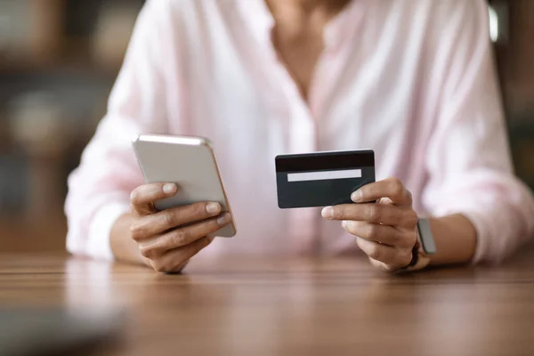 Cropped of woman in pink blouse sitting at desk, holding modern cell phone and plastic credit card, unrecognizable lady shopping online or paying bills on Internet. Online banking concept