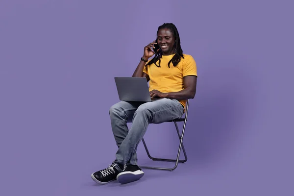 明るく若いアフリカ系アメリカ人男性と長い編組でカジュアルな衣装を着たフリーランスの椅子に座って 現代のラップトップで作業し 顧客と電話をかけ 紫色のスタジオの背景 コピースペース — ストック写真