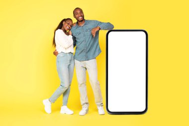 Boş Ekran Reklamcılığı Uygulaması Olan Büyük Telefonun Yakınındaki Siyah Çift Ya da Sarı Arkaplandaki Kamera Pozuna Gülümseyen Mobil Teklif. Teknoloji ve Gadget Kavramı. Stüdyo Çekimi, Model